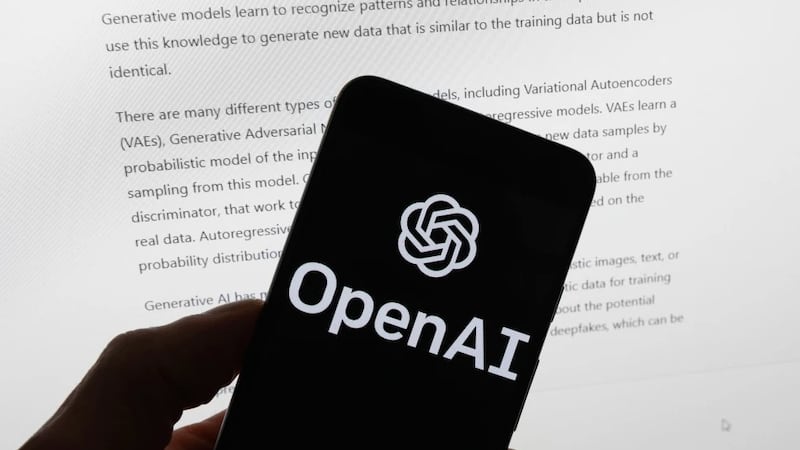 The OpenAI logo is displayed on a mobile phone in front of a computer screen with output from...